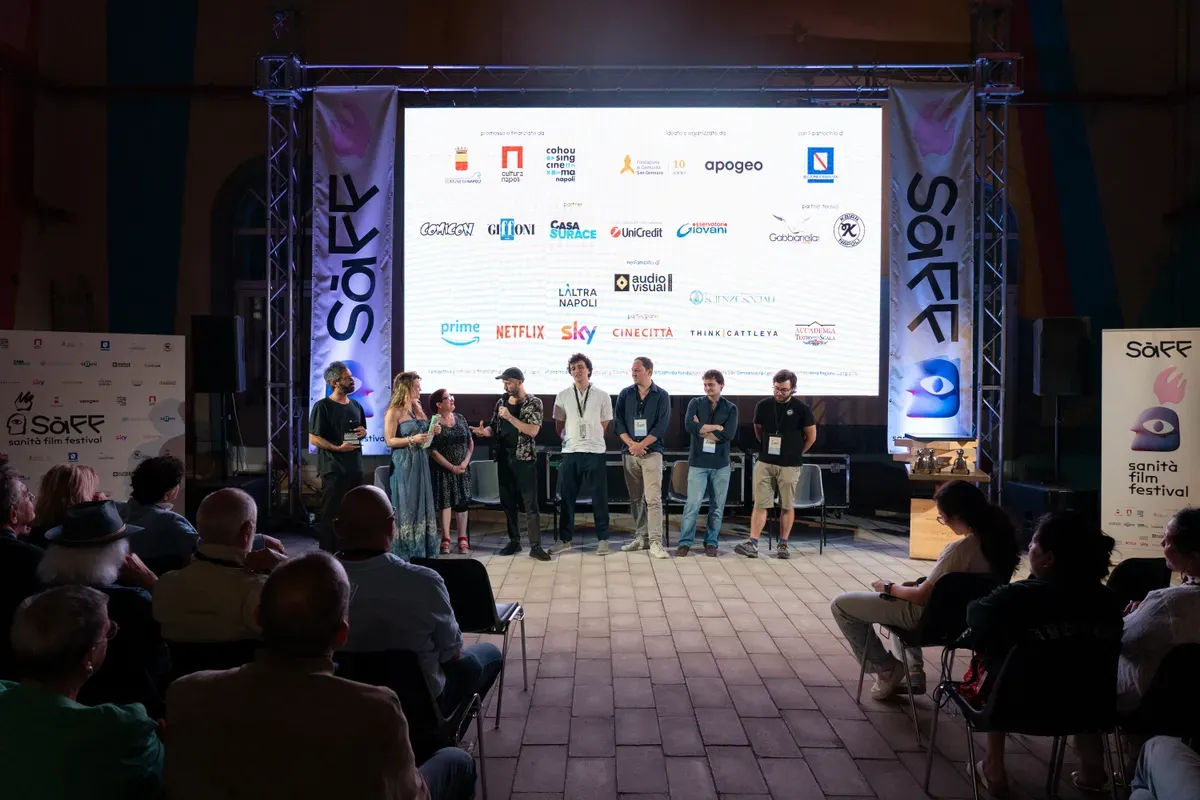 Otto persone in piedi sul palco davanti a uno schermo che mostra i loghi dei promotori e dei partner, con il pubblico seduto di spalle in primo piano