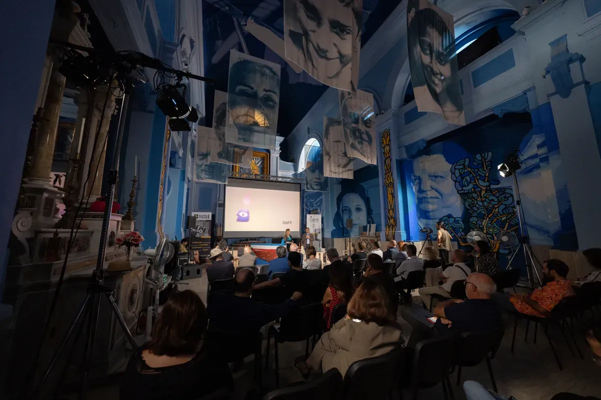 La sala di una chiesa sconsacrata con teli stampati appesi in alto che ritraggono volti, il pubblico seduto in file e uno schermo acceso con il logo a forma di occhio