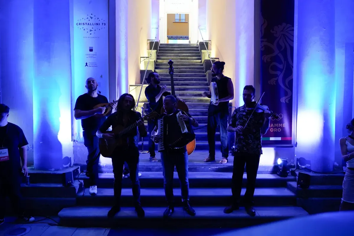 Sei musicisti suonano su una scalinata illuminata di blu, con mandolino, chitarra, contrabbasso, tamburello, fisarmonica e violino, davanti a un manifesto con la scritta Cristallini 73