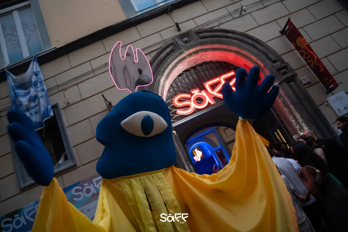 Un grande pupazzo azzurro con un solo occhio e un mantello giallo alza le braccia davanti al portale del festival, illuminato da una scritta SàFF al neon rosso