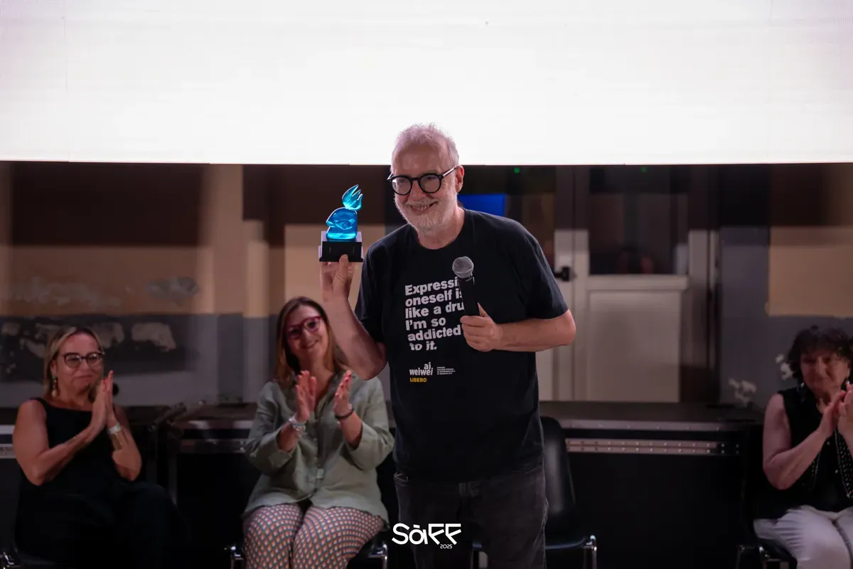 Un uomo con barba bianca e occhiali tondi sorride sul palco mostrando un premio di vetro azzurro a forma di fiamma, mentre tre donne sedute dietro di lui applaudono
