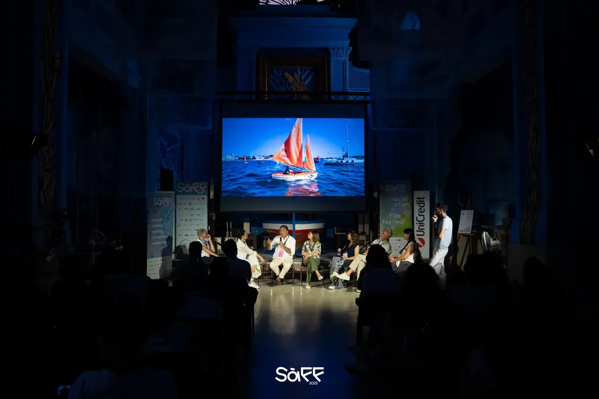 Sala al buio durante una proiezione: sullo schermo una barca a vela con le vele arancioni, e sotto un gruppo di relatori seduti davanti al pubblico in controluce
