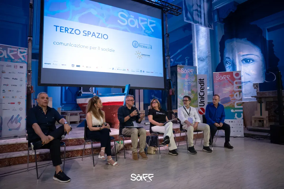 Sei relatori seduti sul palco sotto uno schermo con la scritta Terzo Spazio, comunicazione per il sociale, in una sala decorata da un grande murale azzurro con un volto