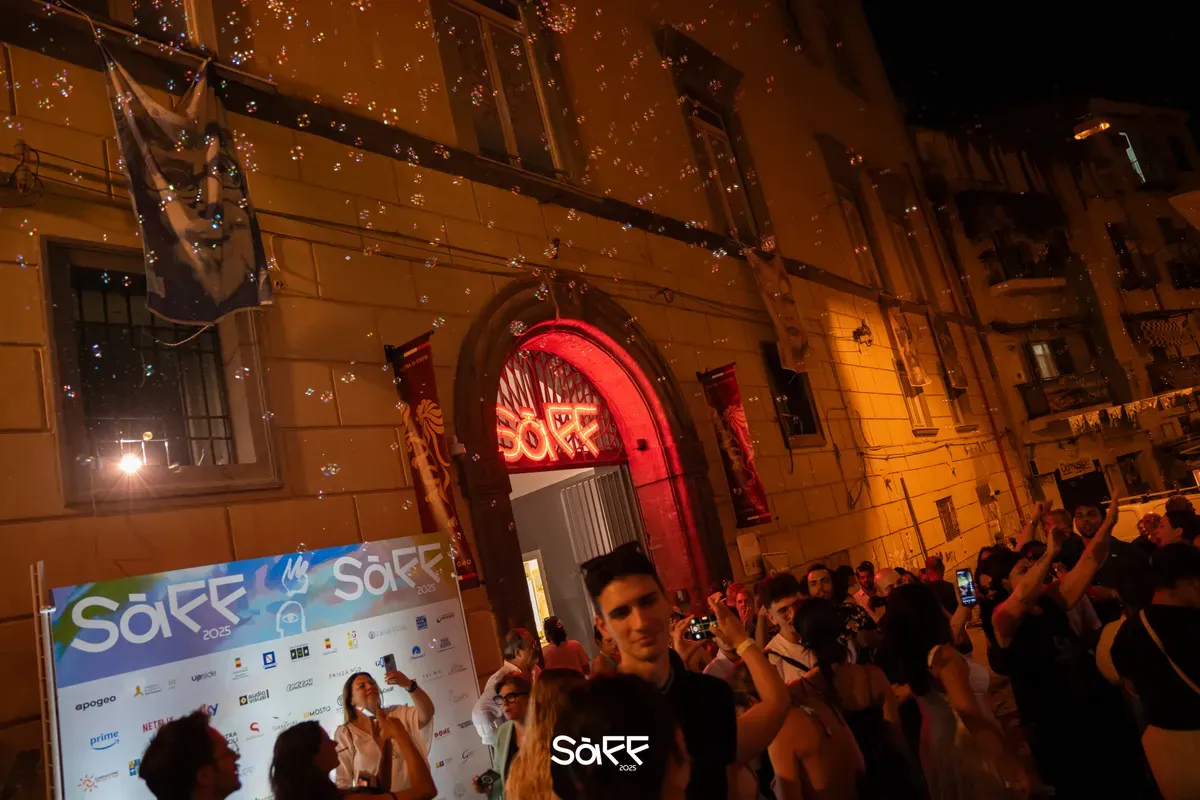 Folla in strada di sera davanti al portale illuminato dal neon rosso del festival, tra bolle di sapone e un pannello con i loghi degli sponsor