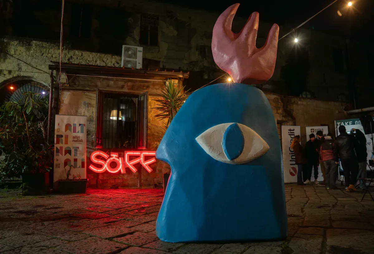 Una grande scultura azzurra a forma di testa di profilo con un solo occhio, in un cortile di pietra di sera, accanto a una scritta SàFF al neon rosso