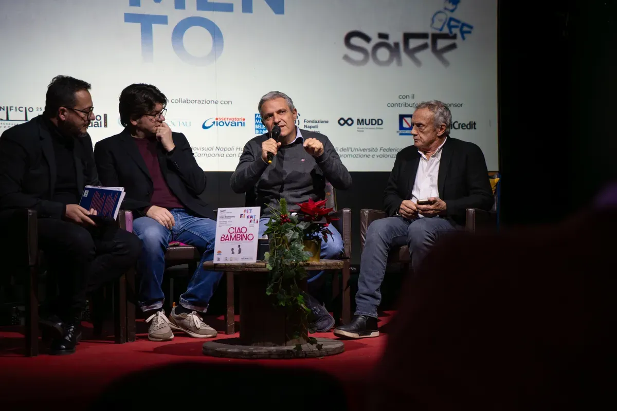 Quattro uomini seduti in poltrona discutono; al centro uno parla al microfono, sul tavolino il libro Ciao Bambino accanto a una stella di Natale